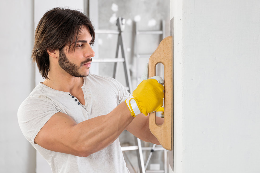 Handsome male plasterer with straight, dark hair and designer facial hair. He is holding a plastering tool against the wall and concentrating. He is wearing a cream coloured t-shirt and displaying his very large bicep.