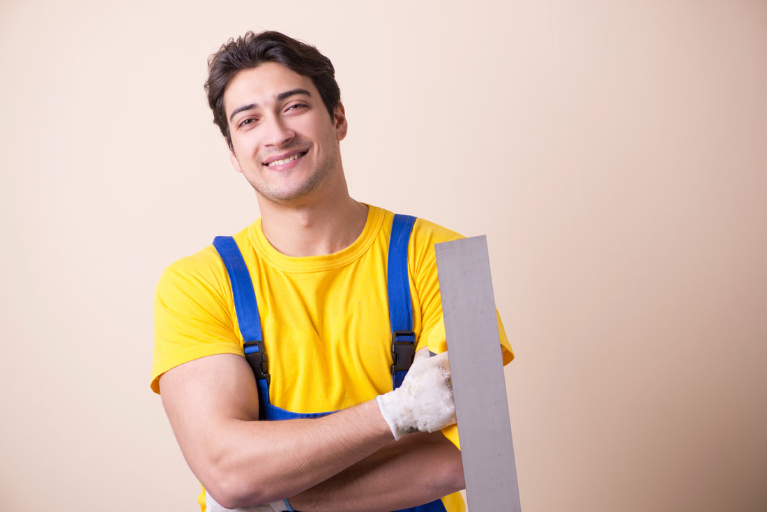 Friendly, handsome, male plasterer wearing a yellow t-shirt and blue dungarees. He has dark hair and is holding a plastering tool in a gloved hand. His arms are folded across his body and he has large biceps on display.