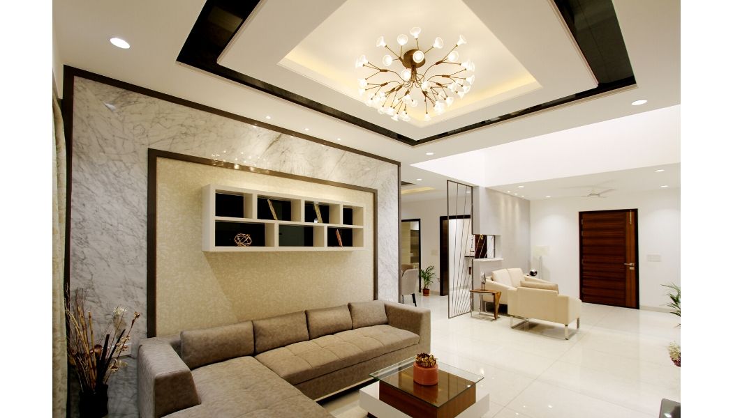 Opulent reception area of a luxury apartment. Large glossy cream floor tiles and dark teak wooden entry door. Beige corner sofa with marble effect wall and elevated bookcase. Glass top coffee table. Magnolia walls and ceiling with black gloss frame around a centrepiece chandelier.