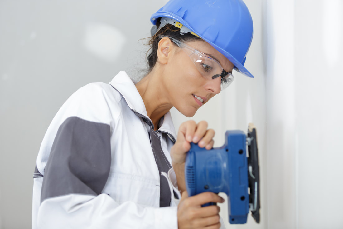 lady plasterer wearing blue hard hat, protective goggles and white and grey overalls. She is holding a blue sanding tool.