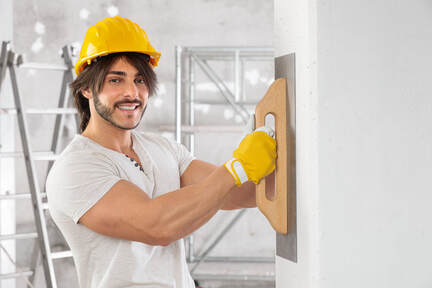 Handsome male plasterer wearing hard hat and smoothing walls. He has designer facial hair, large biceps and a lovely smile.